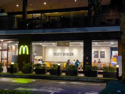 Thai and chinese adults in McDonald's shop in Bangkok at deep night. View from sidewalk to main windows. Restaurant is seated at Ratchadaphisek Rd in area Huai Khwang - Suttisan at main road. Some people are sitting inside. They are eating and using mobile phones. At wall inside are letters TASTY BURGER. Illuminated yellow M logo is at left side near entrance.