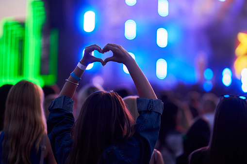Love symbol gesture, fans celebrate artist, youth culture, happy summer vibe in concert. Person forms heart shape with hands at lively outdoor music festival, blurred stage lights, crowd enjoys event.