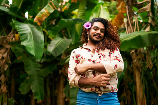 Confident smile, celebrates inclusivity, pride, LGBT identity amidst natures embrace. South Asian gay man with floral shirt, blue jeans stands in tropical foliage, purple flower in hair.