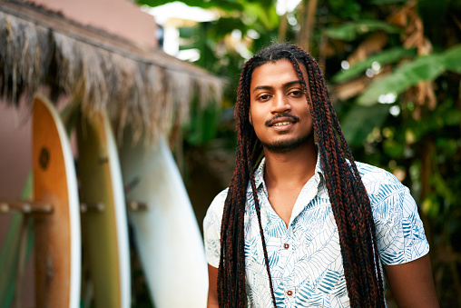 Surfboards align in background. Confident coach with dreadlocks stands at surf camp. Instructor with warm smile invites for lessons. Black surfer promotes sport, inclusion tropical retreat.