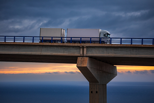 Three-axle truck with trailer driving along a viaduct, with the sea on the horizon.