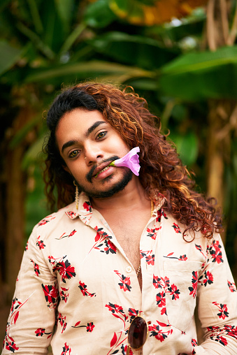Expresses freedom, LGBT pride, gender fluidity. Gay man with curly hair and floral shirt stands in garden, purple flower in mouth. Relaxed, confident pose in natural setting. Symbolizes love, spring.