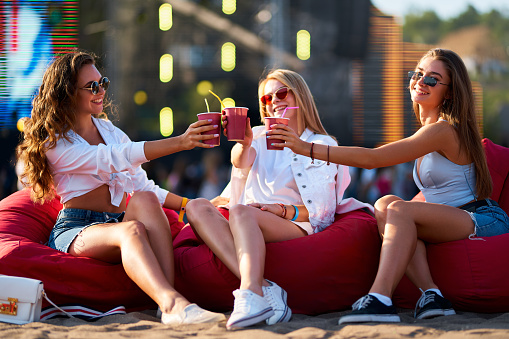 Three carefree friends in summer outfits, sunglasses clink cups, relax on coast. Group of happy young females sit on red beanbags at sunny seaside music festival, cheers with drinks, chill vibes.