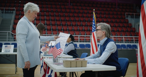 Elderly woman talks with polling officer and takes vote bulletin. American citizens come to vote in polling station. Political races of US presidential candidates. National Election Day. Civic duty.