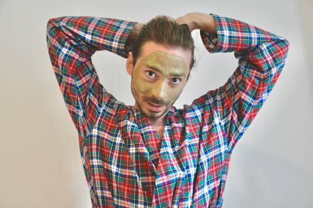 Young man with a clay face mask, skin care concept in plaid robe  while tying up his hair on white background with copy space