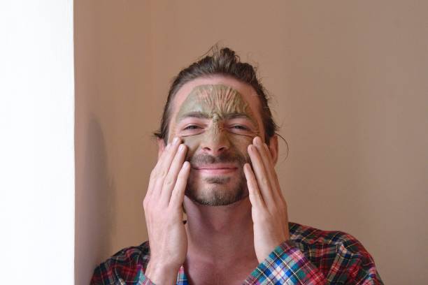 Young man with a clay face mask, skin care concept in plaid robe grimaces in annoyance