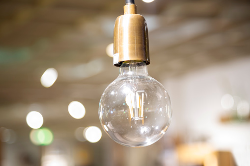 Incandescent light bulb with tungsten filaments in golden socket hanging down from a ceiling against bright bokeh lights. Shallow depth of filed.