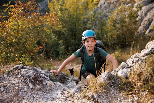 Boy with helmet and backpack is climbing on rock while hiking in the mountains