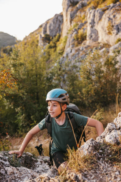 Boy with helmet and backpack is climbing on rock while hiking in the mountains