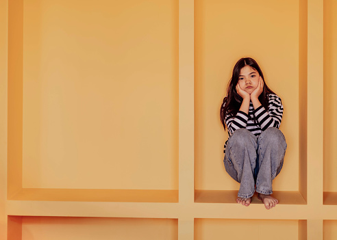 Asian girl sits within geometric structure, her pose indicating a sense of restriction or contemplation, themes of confinement and introspection