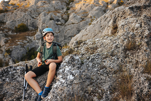 Active boy with trekking poles and backpack sitting on rock and resting of hiking on rocky mountain