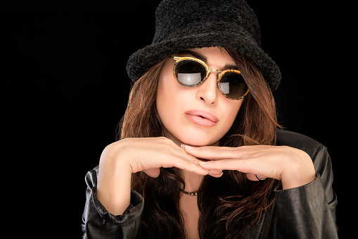 Fashionable beautiful young woman wearing stylish modern sunglasses and trendy autumn outfit resting her chin on her hands as she looks pensively at the camera. isolated on black