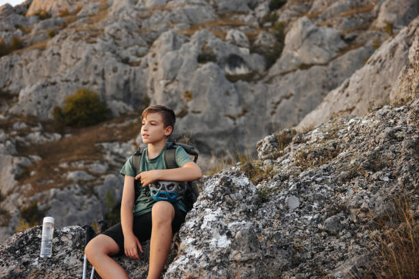 Active boy with trekking poles and backpack sitting on rock and resting of hiking on rocky mountain
