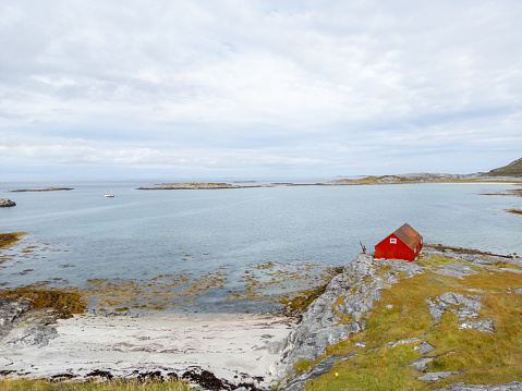A red cabin on the rocky shore of a calm bay. The vibrant red color of the cabin stands out against the green grass, blue sky, and tranquil waters. The sky is scattered with clouds, creating a sense of serenity and solitude in a remote natural setting.