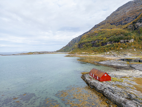 A serene landscape with a solitary red cabin nestled on the rocky shore of a tranquil bay. The cabin's vivid red color contrasts beautifully with the subtle greens of the grassy terrain, the pale blues of the sky, and the calm waters at the base of a mountain. Clouds softly scatter the sky, suggesting a peaceful, remote retreat in a natural setting.