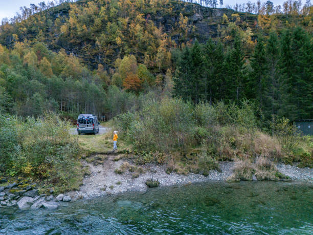 An adventurous scene with an off-road vehicle parked by a mountain river's edge, amidst an autumnal forest setting. A person in a yellow top stands beside the vehicle, ready to explore the wilderness. The backdrop of a steep cliff with trees displaying a medley of fall colors—from vibrant oranges to deep greens—creates a striking contrast against the calm, clear river.