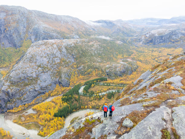 Two hikers, uniformly dressed in blue jackets, jeans and boots, each carrying red backpacks, stand on a mountain peak, looking down at a valley filled with the golden colors of autumn.  The male hiker has a golden yellow head warmer on to match the hints of yellow on his jacket. The view includes rivers, forests, and rugged mountains, all covered in a gentle mist. This scene beautifully represents the bond between humans and the vast wilderness, showcasing the beauty of nature and the sense of adventure.