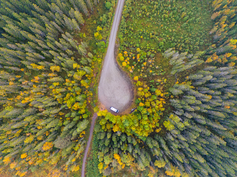 Looking straight down from above, there's a spot where the road bends like a teardrop in the woods. It looks welcoming for those who love to travel and explore. The trees are thick but you can see bright yellow and green leaves of early fall peeking through. A camper van sits where the roads meet, making the place look even more ready for adventure. The way the clearing is shaped and how the two roads come together makes it an interesting and exciting picture, full of the promise of adventure in nature.