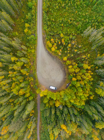 Looking straight down from above, there's a spot where the road bends like a teardrop in the woods. It looks welcoming for those who love to travel and explore. The trees are thick but you can see bright yellow and green leaves of early fall peeking through. A camper van sits where the roads meet, making the place look even more ready for adventure. The way the clearing is shaped and how the two roads come together makes it an interesting and exciting picture, full of the promise of adventure in nature.