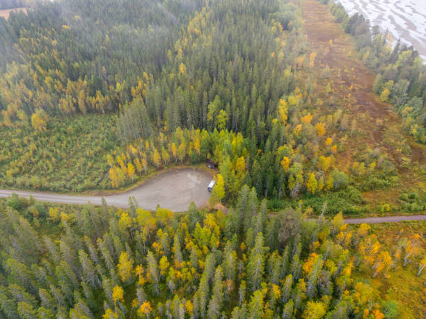 Aerial view of a quiet spot by a lake, surrounded by a soft fog that highlights the peacefulness of the area. The forest around it, hinting at autumn with spots of yellow among the green, encloses a little open space where a vehicle has stopped. The big, calm lake blends into the distance, with patterns on its surface from the water pulling back.
