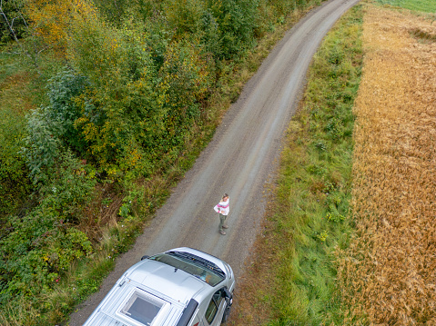 A person standing on a tranquil country road, bordered by the rich contrast of vegetation and harvested fields. A camper van parked nearby indicates a break in the journey, suggesting a moment for reflection or enjoyment of the natural landscape. Subtle signs of autumn appear in the changing colors of the tree leaves, highlighting the seasonal transition.