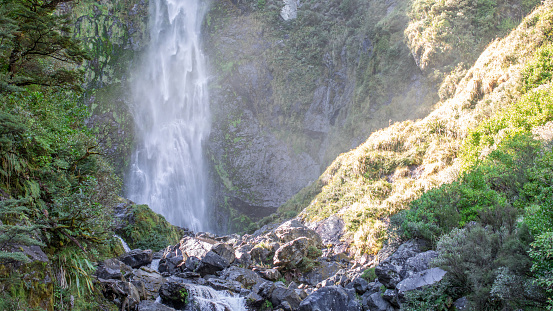 Sunlit waterfall cascades through lush NZ foliage, a serene Arthur's Pass gem. Travel