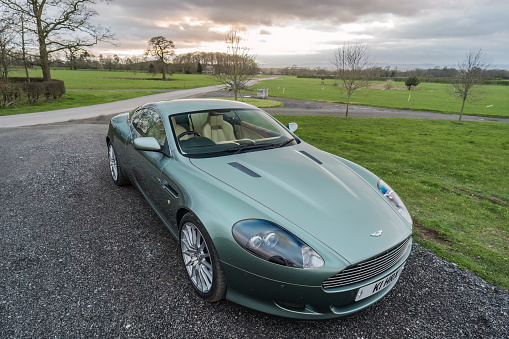 Chester, Cheshire, England, March 15th 2024. A pale green Aston Martin DB9 is parked on a gravel driveway, with countryside and trees in the backdrop.