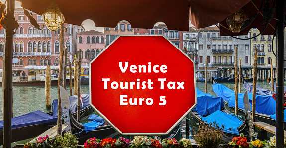 Bold red sign announcing a Venice Tourist Tax of Euro 5 per day is prominently displayed in the foreground, with the picturesque scenery of Venice's iconic canals and moored gondolas in the background
