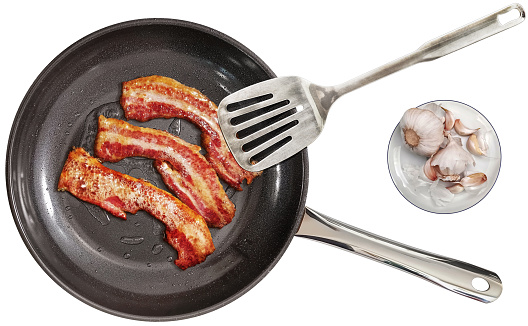 Traditional fried juicy crunchy gourmet bacon rashers in a large new modern heavy duty black frying pan, with non-slip ceramic double coated inner surface, equipped with stainless steel spatula, and pair of Garlic bulbs with few detached cloves on white rimmed porcelain plate set beside, isolated on white background, viewed directly above, high resolution stock image.