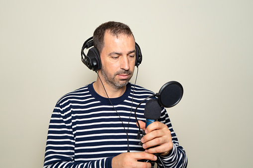 Bearded Hispanic man in his 40s wearing a striped sweater looking confused while inspecting a microphone and headphones, isolated on beige background