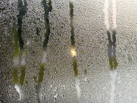 Blurred headlight of a passing car on a suburban street seen through one of several streaks on a window obscured by raindrops early on a spring morning, for motifs of weather and visibility
