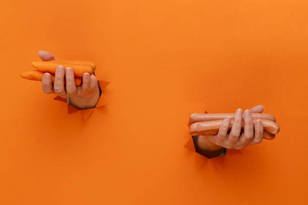 Concept of choice: fresh carrots in one hand, sausages in the other hand. Orange background