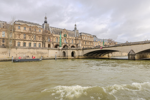 Paris, France - December 29, 2023:  Musee du Louvre and Pont du Carrousel as viewed from a Seine River cruise boat on a cloudy day as it's about to pass underneath the bridge.  HDR encoded