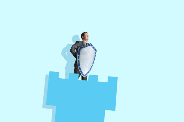 A businessman stand with a medieval shield as he stands in a castle turret. Isolated against a light blue background.