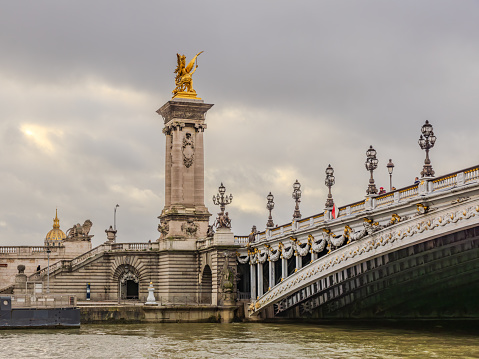 Paris, France - December 29, 2023:  The beautiful Pont Alexandre III as viewed from a Seine River cruise boat on a gloomy day, with the golden dome of Hotel Des Invalides visible on the left.  HDR encoded