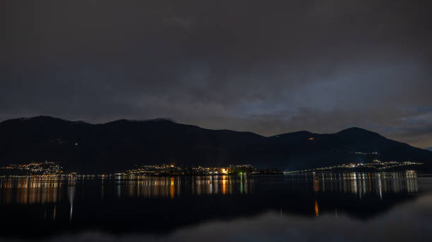 Long exposure captures the serene beauty of Lake Bourget under a vast, star-filled night sky with large copy space. The distant lights of Aix-les-Bains twinkle along the shoreline, creating a gentle glow against the dark silhouette of the surrounding mountains. This photo was taken from Grand Colombier summit in Bugey mountains, in Ain department in Auvergne-Rhone-Alpes region in France, during a summer night with clear starry sky.