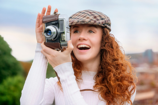 enjoying smiling young redhead lady taking pictures on vintage camera in city. Hobby and leisure concept