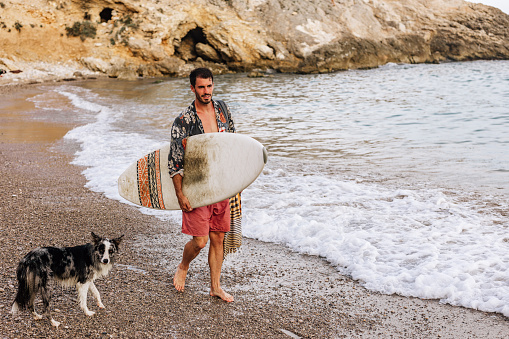 Young male surfer and his loyal dog enjoying the sun on a sandy beach and preparing their surfboard for a big waves. Man listening to music with a headphones. Perfect sunny day.