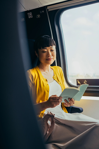 A middle-aged Asian woman enjoys reading a novel while traveling in a quiet train compartment, reflecting calm and leisure during transit.