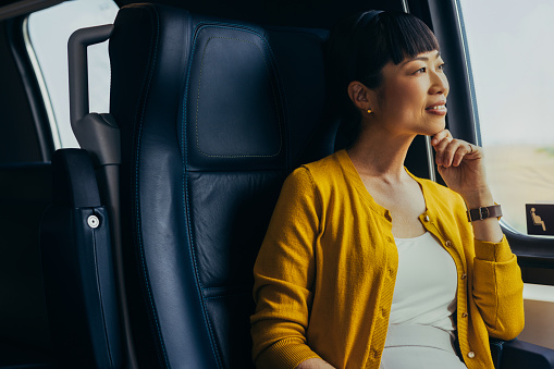 An elegant Asian woman dressed in a yellow cardigan gazing out the window during a car ride, reflecting a serene and thoughtful mood. The interior suggests luxury and comfort.