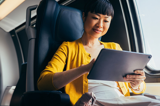 A professional mature Asian businesswoman uses a digital tablet while seated in a comfortable airplane chair. The scene captures a moment of focused productivity during a flight.