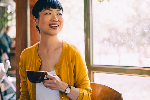 A cheerful Asian woman in a yellow cardigan enjoys her coffee at a sunny café, embodying comfort and contentment. Ideal for lifestyle and leisure themes.