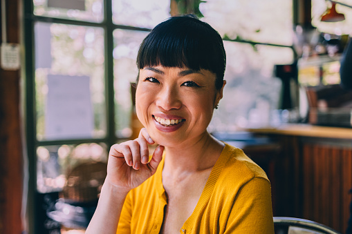 Portrait of a smiling Asian woman wearing a bright yellow sweater, sitting comfortably in a rustic café setting with natural light pouring in.