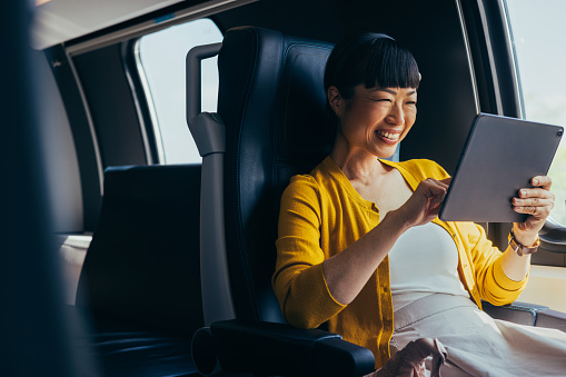 Asian businesswoman smiles joyfully while using a tablet in a luxurious car, portraying comfort and productivity during travel. Her yellow cardigan and casual smart attire enhance the warm ambiance.