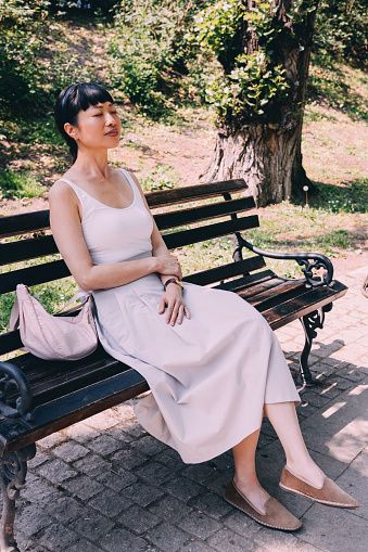 A poised woman enjoys a moment of tranquility on a sunlit park bench, exuding elegance and calm amidst lush greenery. A perfect blend of relaxation and style.