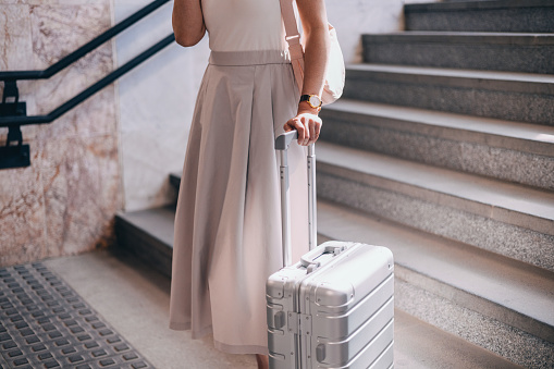 A stylish, well-dressed woman stands on a staircase, holding a suitcase, seemingly ready for travel. The image captures a sense of elegance and anticipation.