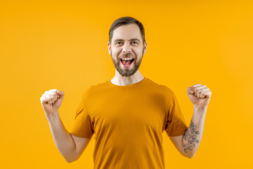 Studio portrait of bearded attractive joyful young man clenching his fists in winners gesture while posing over yellow background.