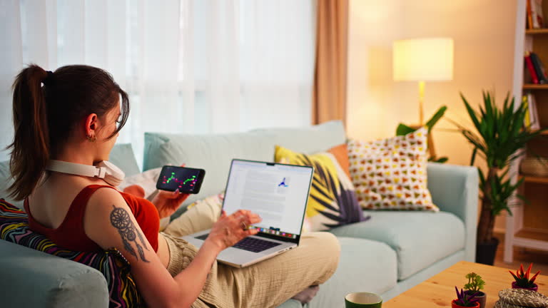 Young Businesswoman sits on the couch with her feet up, coffee cup in hand, checking the Bitcoin or exchange price chart on a digital exchange on her cell phone while doing research on a laptop monitor