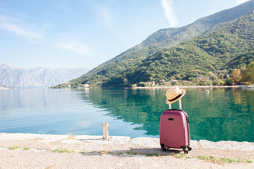 Pink suitcase with straw hat on sea beach. Concept of travel, vacation, female tourism, trip, journey, adventure. Nature background of amazing view with blue lake, coast, green mountains.