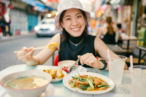 Singapore vacation asian woman smiles teasingly as she sits with her friend at a table in a sidewalk cafe in Singapore's Chinatown. eating rice with asian street menu fried tufu and grill pork with vegetable